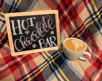 May include: A chalkboard sign with white lettering that reads "HOT Chocolate BAR" with snowflake designs. A white mug with a brown liquid and a heart-shaped design in the foam sits on a red and white plaid blanket.