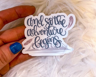 May include: A white sticker with the text "and so the adventure begins" in black cursive lettering. The sticker also features a mountain range and two feathers in a light purple color.