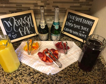 May include: A mimosa bar setup with two bottles of champagne, orange juice, cranberry juice, and fresh fruit garnishes. The sign reads "Mimosa Bar" and "Pour some bubbly, splash some juice, garnish with fruit".