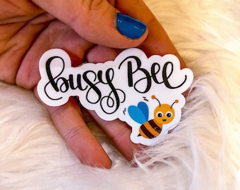 May include: A white sticker with the text "busy Bee" in black cursive font. A cartoon bee with yellow and black stripes, blue wings, and a smiling face is below the text.