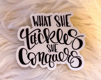 May include: White sticker with black lettering that reads "What she tackles she conquers".