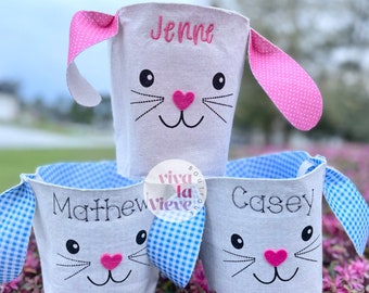 Puede incluir: Tres cestas de Pascua blancas con forma de conejo con orejas de vichy rosa y azul. Las cestas están personalizadas con los nombres "Jenne", "Matthew" y "Casey".