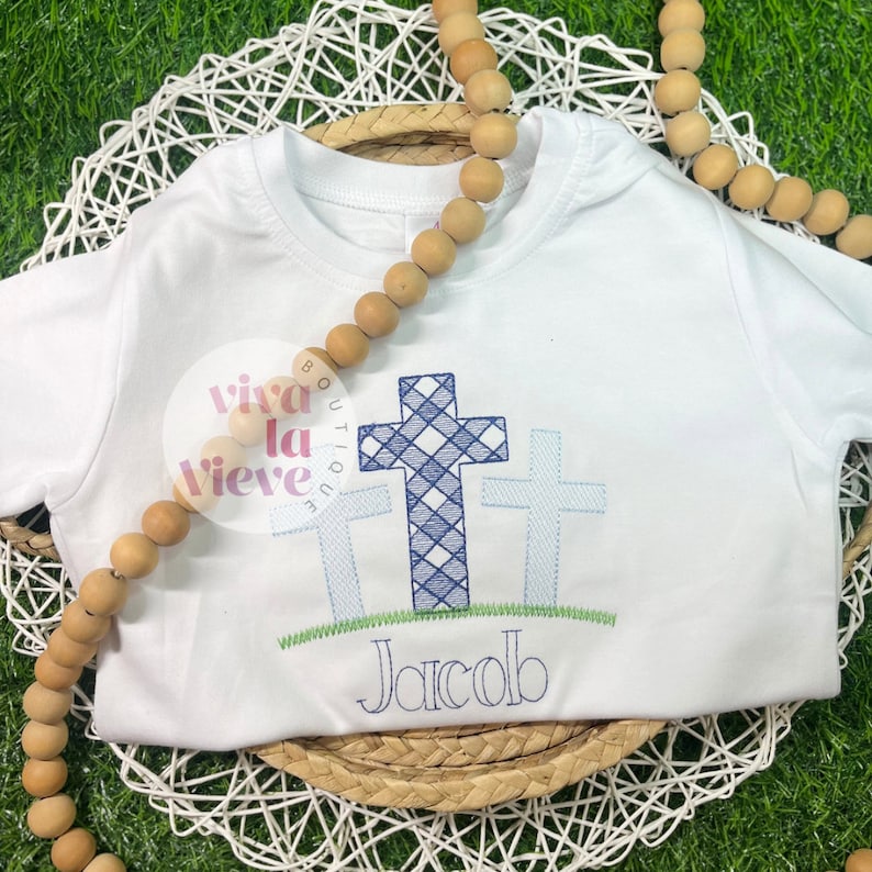 Pode incluir: Um macac&atilde;o branco para beb&ecirc; com uma cruz xadrez azul e branca e duas cruzes menores. O nome "Jacob" est&aacute; bordado abaixo das cruces.