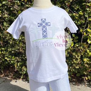 Pode incluir: Camiseta branca com tr&ecirc;s cruzes xadrez azuis e brancas e o texto "Matthew" bordado na frente. A camiseta est&aacute; em um manequim vestindo shorts listrados azuis e brancos.