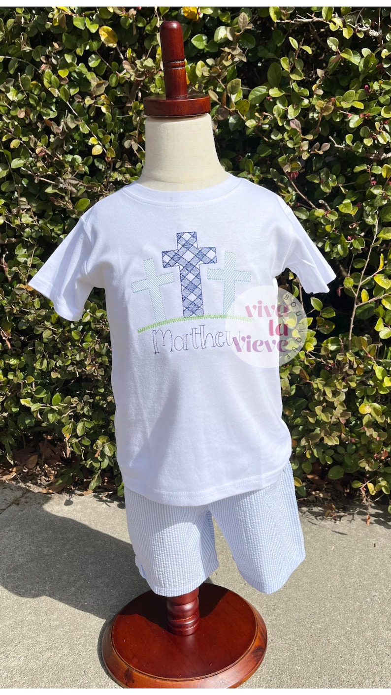 Pode incluir: Camiseta branca com tr&ecirc;s cruzes xadrez azuis e brancas e o texto "Matthew" bordado na frente. A camiseta est&aacute; em um manequim vestindo shorts listrados azuis e brancos.