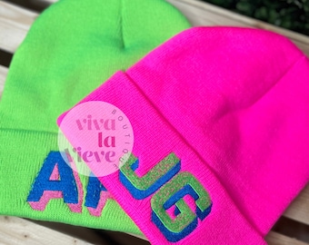 May include: Two knit beanies, one green and one pink, with embroidered text. The green beanie has the word "Alive" embroidered in blue and pink. The pink beanie has the word "G.L.O.W" embroidered in blue and green.