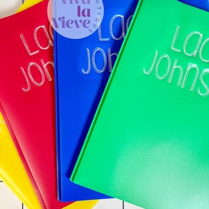 May include: A stack of colorful notebooks in yellow, red, blue, and green. Each notebook has the name "Lacy Johns" written in white script. The notebooks are arranged on a white grid surface with a keyboard in the background.