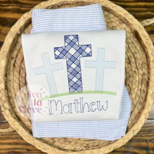Pode incluir: Macac&atilde;o de beb&eacute; branco bordado com tr&ecirc;s cruces azuis e o nome "Matthew" por baixo. As cruces s&atilde;o bordadas com um padr&atilde;o xadrez azul e branco.