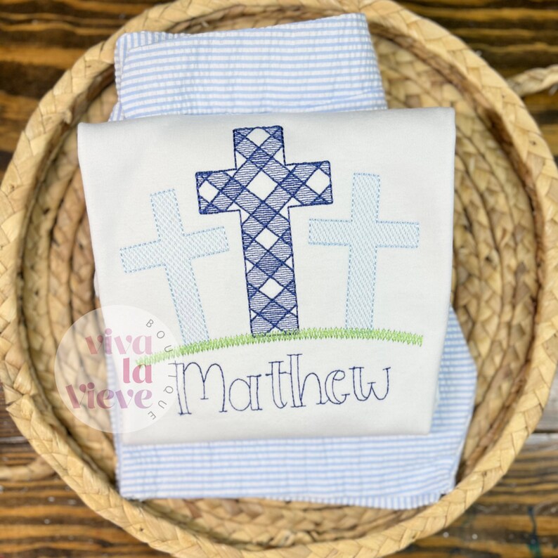 Pode incluir: Macac&atilde;o de beb&eacute; branco bordado com tr&ecirc;s cruces azuis e o nome "Matthew" por baixo. As cruces s&atilde;o bordadas com um padr&atilde;o xadrez azul e branco.