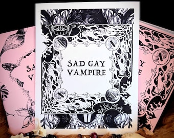 May include: A stack of three art prints. The central print is white with black illustrations and the text "SAD GAY VAMPIRE." Two pink prints with black illustrations are on either side. The prints are on a brown fur-like surface with small bones and beads.