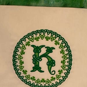 以下が含まれることがあります： 緑色の刺繍が施された白い布。ケルトの結び目のデザインで、縁にはクローバーが飾られ、中央にはKの文字が刺繍されています。布の下部には「TDD Embroidery」の文字があります。