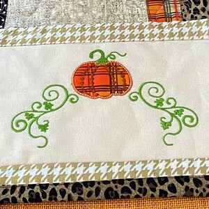 Könnte beinhalten: Ein weißer Tischläufer mit Patchwork-Design aus orangefarbenen, schwarzen und grünen Quadraten. Der Läufer hat einen braunen und schwarzen Leopardenmuster-Rand und einen Fransenrand. Die Mitte des Läufers zeigt einen gestickten Kürbis mit grünen Ranken.