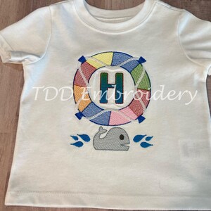 Può includere: T-shirt bianca a maniche corte con un design ricamato. Il design presenta un salvagente colorato con la lettera "H" al centro, una balena grigia e spruzzi d'acqua blu. Il testo "TDD Embroidery" è sotto il colletto.