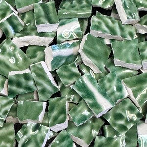 May include: A collection of irregularly shaped, broken ceramic tiles. The tiles are primarily a deep green color with white and lighter green marbling. The edges of the tiles are a rough, off-white color, suggesting a mosaic or craft project.