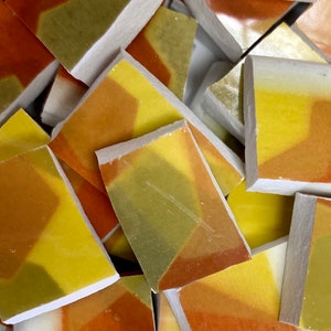 May include: A close-up shot of a pile of small, rectangular mosaic tiles. The tiles feature a geometric design with shades of yellow, orange, and green. The edges are white, and the tiles appear to be made of ceramic or a similar material.