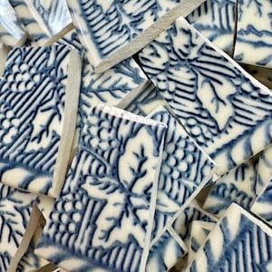 May include: A collection of broken ceramic tiles with a blue and white floral pattern. The tiles are rectangular and feature a detailed design of leaves and berries. The broken edges reveal a light gray interior.