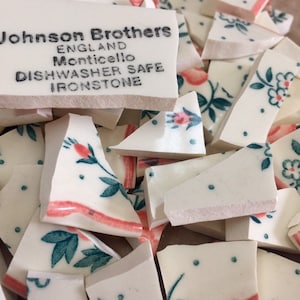 May include: A collection of broken white ceramic pieces with floral patterns in pink and teal. The pieces are from Johnson Brothers, England, Monticello, Dishwasher Safe, Ironstone. The fragments vary in size and shape, creating a scattered arrangement.