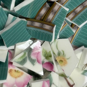 May include: A collection of broken ceramic pieces with various designs. Some pieces feature teal and white stripes, while others display floral patterns in pink, yellow, and green. The fragments vary in size and shape, creating a mosaic-like appearance.