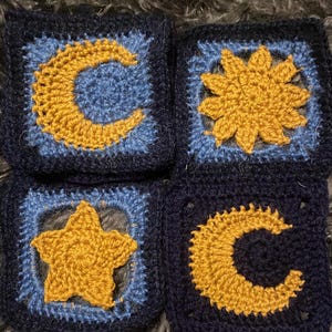 May include: Four crocheted squares with different celestial designs. The squares are navy blue with a light blue border and feature a yellow crescent moon, a yellow sun, a yellow star, and a yellow crescent moon.