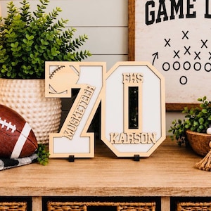 May include: A wooden tabletop sign featuring the number 70 with a football cutout, the text Musketeer, EHS, and Karson. The sign has a light wood frame and a white background, standing on small black bases on a wooden surface.