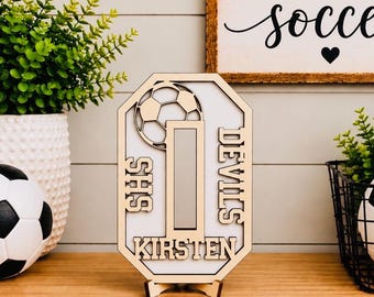 May include: A light-colored wooden sign shaped like an octagon featuring a cutout of the number 1, a soccer ball graphic, and the text SHS, DEVILS, and KIRSTEN. The sign is displayed on a small wooden stand on a flat surface.