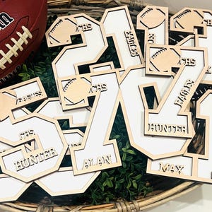 May include: A collection of laser-cut wooden number cutouts featuring white inserts. Each number is decorated with a small football icon and engraved text including names like Hunter, Alan, and Max, along with the acronyms CHS and Eagles.