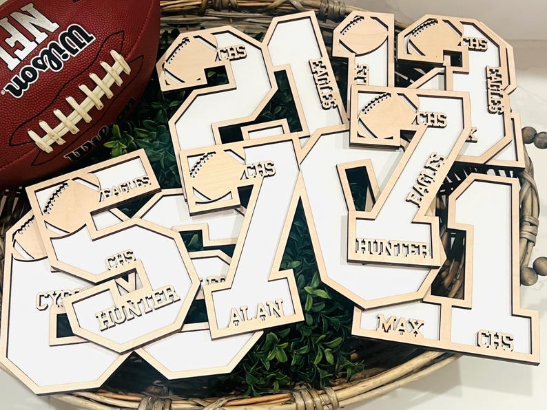 May include: A collection of laser-cut wooden number cutouts featuring white inserts. Each number is decorated with a small football icon and engraved text including names like Hunter, Alan, and Max, along with the acronyms CHS and Eagles.