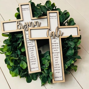 May include: Two wooden crosses with the names "Brynn" and "Cali" engraved on them. The crosses are decorated with the text "Baptized" and a date, and a Bible verse from Joshua 1:9. The crosses are displayed on a green wreath.