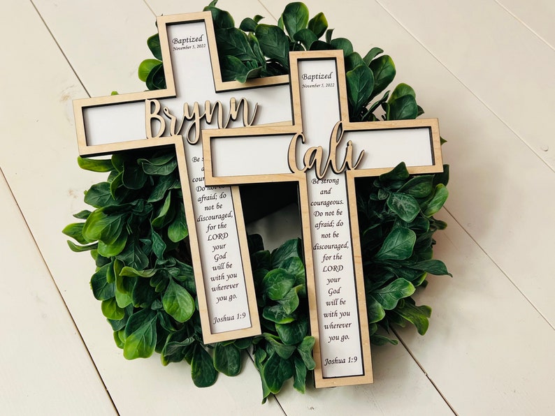 May include: Two wooden crosses with the names "Brynn" and "Cali" engraved on them. The crosses are decorated with the text "Baptized" and a date, and a Bible verse from Joshua 1:9. The crosses are displayed on a green wreath.