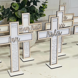 May include: A set of wooden crosses with personalized names and a scripture verse. The crosses are painted white and have a natural wood finish. The crosses are standing on a white surface.