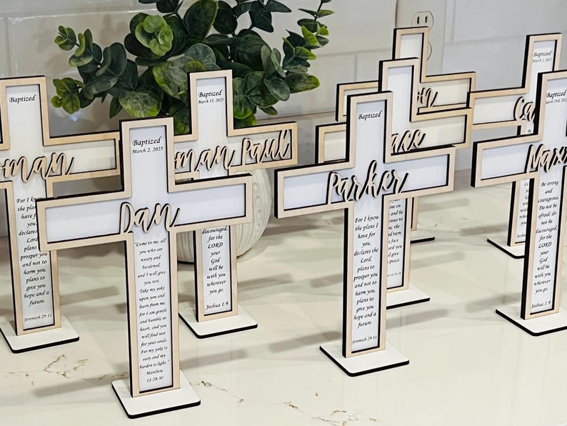 May include: A set of wooden crosses with personalized names and a scripture verse. The crosses are painted white and have a natural wood finish. The crosses are standing on a white surface.