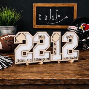 May include: A wooden display sign featuring the numbers 2212 with white inserts. The design includes football icons and the text "COLE", "CASH", "EARLHAM", and "CARDINALS" laser-cut into the wood. The sign stands on a flat surface as a decor piece.