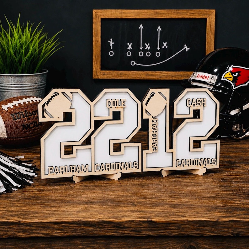 May include: A wooden display sign featuring the numbers 2212 with white inserts. The design includes football icons and the text "COLE", "CASH", "EARLHAM", and "CARDINALS" laser-cut into the wood. The sign stands on a flat surface as a decor piece.