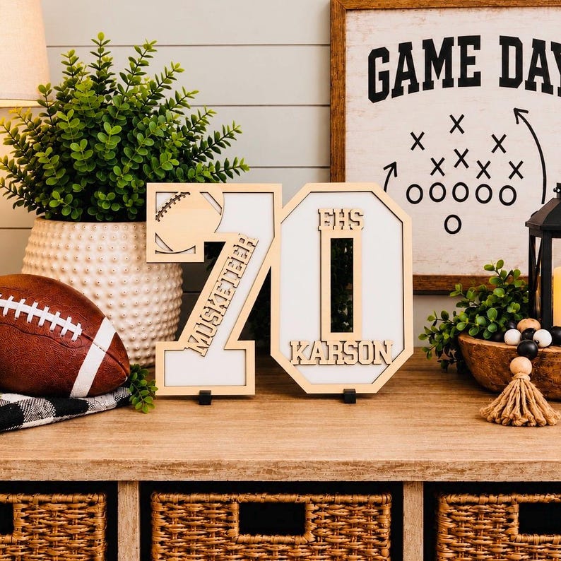 May include: A wooden tabletop sign featuring the number 70 with a football cutout, the text Musketeer, EHS, and Karson. The sign has a light wood frame and a white background, standing on small black bases on a wooden surface.