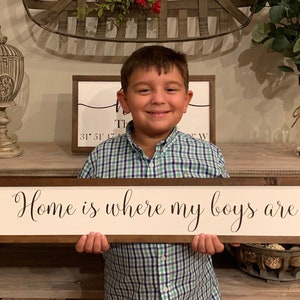 May include: A young boy in a green and blue plaid shirt holds a wooden sign with the text "Home is where my boys are" in black cursive lettering.