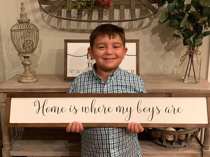 May include: A young boy in a green and blue plaid shirt holds a wooden sign with the text "Home is where my boys are" in black cursive lettering.