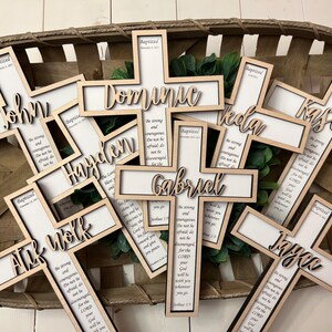 May include: A collection of wooden crosses with names and a bible verse inscribed on them. The crosses are arranged in a basket and are all in a light wood tone.