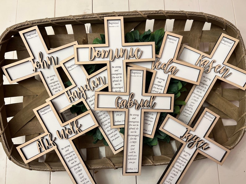 May include: A collection of wooden crosses with names and a bible verse inscribed on them. The crosses are arranged in a basket and are all in a light wood tone.