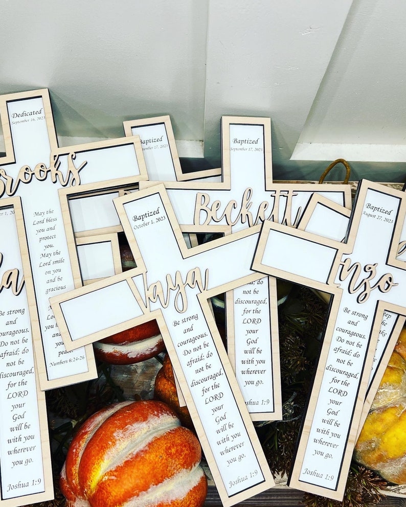 May include: A collection of wooden crosses with personalized names and the text "Be strong and courageous. Do not be afraid; do not be discouraged, for the Lord your God will be with you wherever you go. Joshua 1:9" printed on them.