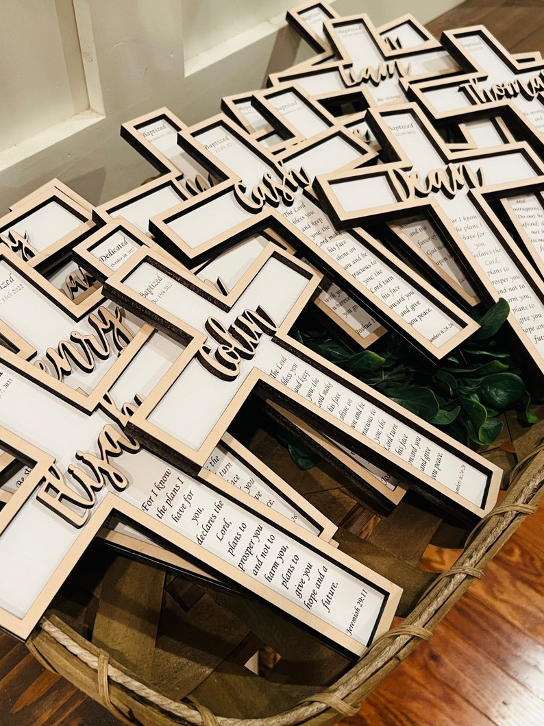 May include: A basket filled with wooden crosses, each with a different name and a Bible verse. The crosses are painted in a light wood color and have a clear acrylic insert for a photo.