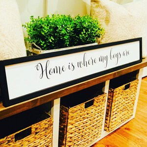May include: A black framed sign with white background reads "Home is where my boys are". The sign is displayed on a wooden shelf with three woven baskets.