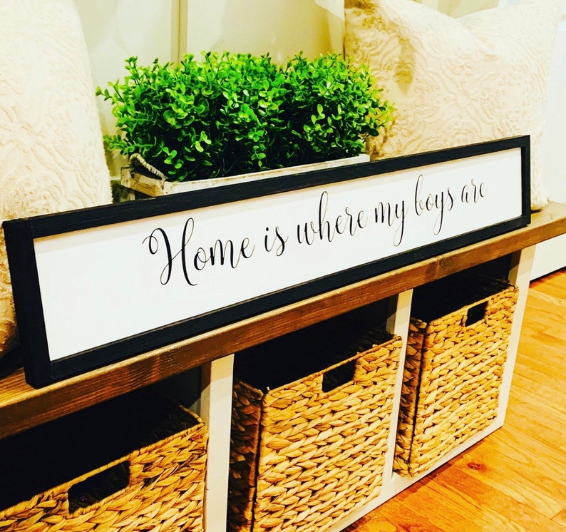 May include: A black framed sign with white background reads "Home is where my boys are". The sign is displayed on a wooden shelf with three woven baskets.