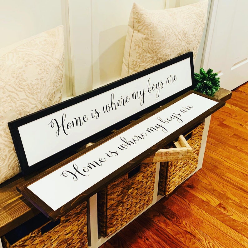 May include: Two wooden signs with white backgrounds and black lettering. The top sign reads "Home is where my boys are". The bottom sign reads "Home is where my boys are". The signs are displayed on a wooden shelf with woven baskets.