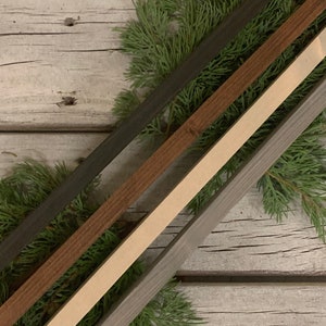 May include: Four wooden strips in different shades of brown, gray, and white, arranged diagonally on a whitewashed wood background. The strips are all the same width and length, and they are partially covered by green pine branches.