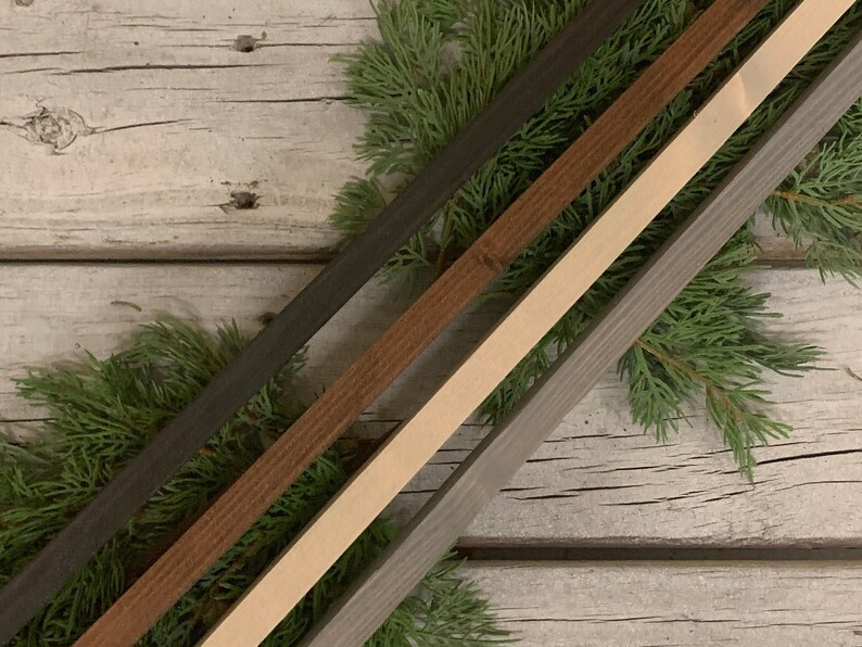 May include: Four wooden strips in different shades of brown, gray, and white, arranged diagonally on a whitewashed wood background. The strips are all the same width and length, and they are partially covered by green pine branches.