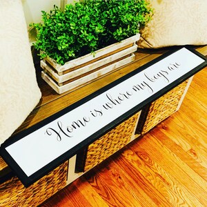 May include: A white wooden sign with black trim and the text "Home is where my boys are" in black cursive lettering. The sign is sitting on a wooden surface with a wicker basket and a potted plant.