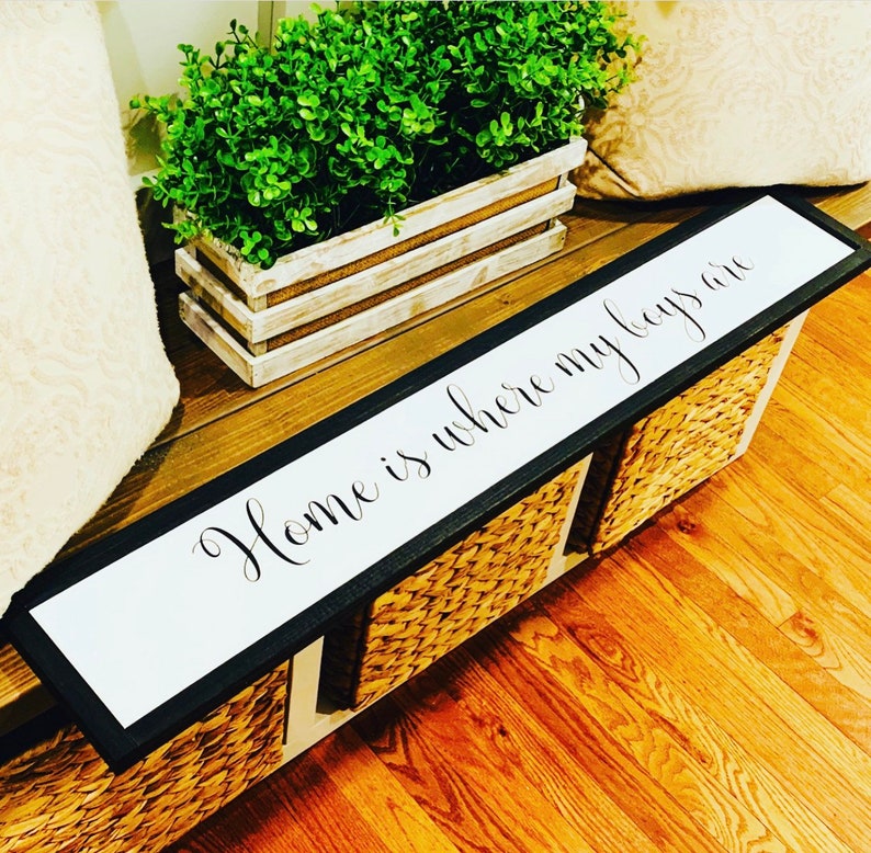 May include: A white wooden sign with black trim and the text "Home is where my boys are" in black cursive lettering. The sign is sitting on a wooden surface with a wicker basket and a potted plant.