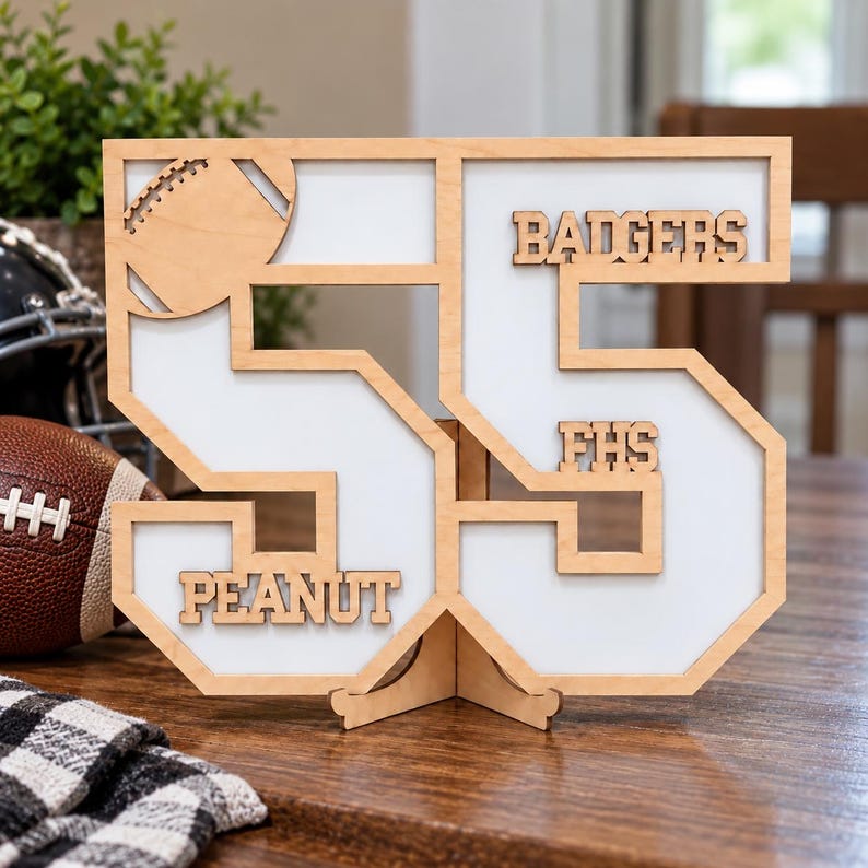 May include: A light-colored wooden cutout of the number 55 with a white backing, featuring a football icon, the text BADGERS, FHS, and PEANUT. The structure is supported by a small wooden stand and displays a layered design with cut-out lettering.