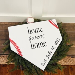 May include: White wooden home plate sign with red stitching and the text "home sweet home" and "Est. July 10, 2023".