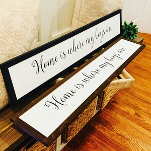 May include: Two wooden signs with white backgrounds and black calligraphy. The top sign reads "Home is where my boys are" and the bottom sign reads "Home is where my boys are".